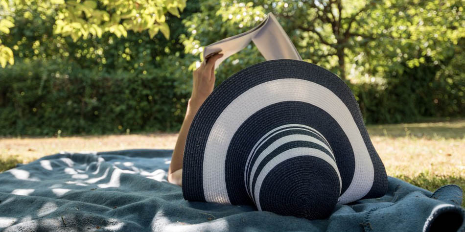 A woman wearing a black and white hat reading a book in the shade of a tree.